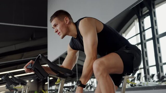 A man is pedaling on an exercise bike at the gym, working out his thigh muscles and strengthening his chest and arm muscles. He is wearing a vest and undershirt