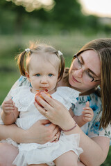 Fototapeta premium Mom feeds a child a peach on a picnic. Mom and girl playing in park at sunset. Mommy and kid spend time together. Portrait of a daughter toddler hugging mother in a field on summer day. Closeup.