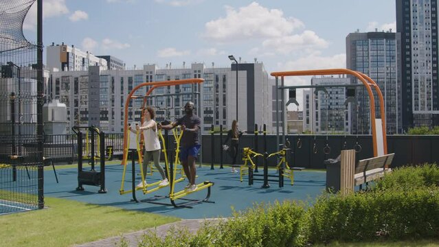 Wide footage of diverse condo neighbours training together on outdoor gym fitness equipment on rooftop area with cityscape on sunny day
