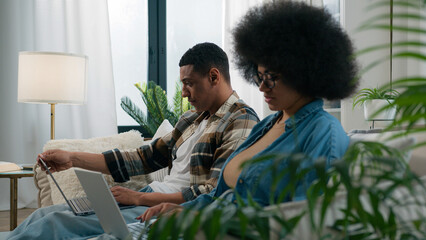 Busy workers freelancers African American man with woman couple at home couch browsing laptops...