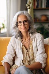 A portrait of a middle-aged woman with gray hair and glasses sitting on a yellow couch