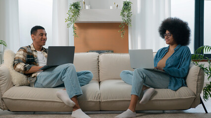 Happy couple relax at couch at home with laptop internet overuse freelancers African American man with woman smiling positive at sofa using typing computers addict pc devices gadgets online gaming app