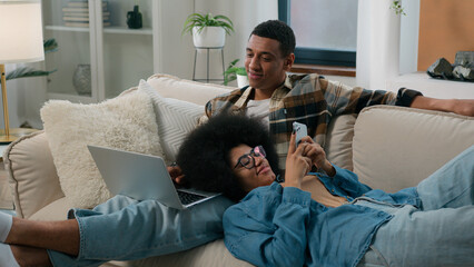 African American couple man with laptop computer sitting on couch home woman chatting smartphone browsing mobile phone laying on male knees girl guy gadget addict internet addiction smiling family