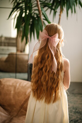 A beautiful girl with long curly red hair and a big bow on her head is standing back at home. Child...