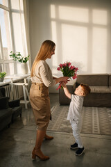Little boy holds big bouquet of tulip and congratulates mother at home. Son gives flowers smiling...