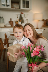Obraz na płótnie Canvas Son hugs smiles mom closeup. Child makes surprise for Mothers Day. Little boy holds, gives bouquet of tulip flowers, congratulates mommy in kitchen. International Women's Day. Happy family holiday.
