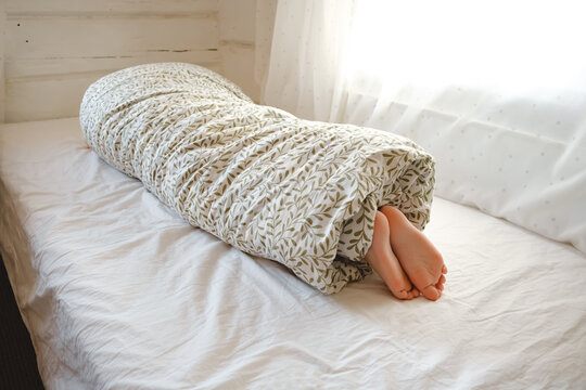 Rolled person. A child wrapped in a blanket with sticking out feet lies on the bed in the bedroom. Sunny morning dream. Psychology comfort. High quality