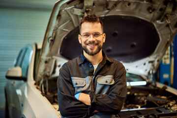 Portrait of a smiling mechanic posing with his hands crossed in front of the opened car hood. - 756407719