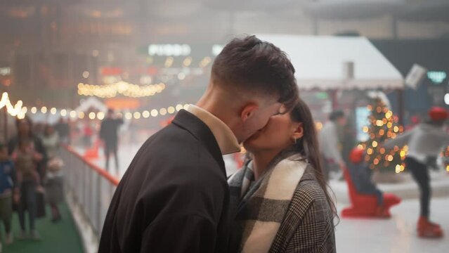 Cheerful young couple in trendy clothes kissing near ice rink. Romantic partners talking and spending leisure time in city on Christmas night. New Year kiss.