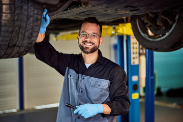 Portrait of a mechanic standing under the car and smiling for the camera, holding a lamp, and checking a tire. - 756408336