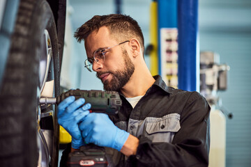 A focused mechanic working in his auto service, using a drill and screw the wheel. - 756408339