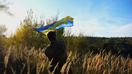 Male military in uniform waving flag of Ukraine at countryside. Young soldier of ukrainian army lifting blue-yellow banner as symbol of victory against russian aggression. Invasion resistance concept. - 756420578
