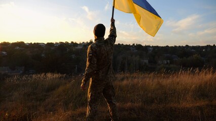 Male soldier stands with raised flag of Ukraine against background of beautiful sunset. Male ukrainian army soldier with a lifted blue-yellow banner in honor of the victory against russian aggression - 756420741