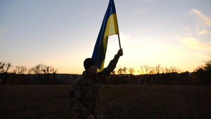 Soldier of ukrainian army going with raised blue-yellow banner on field at sunset. Young male military in uniform walks with flag of Ukraine at meadow. Concept of victory against russian aggression - 756420911