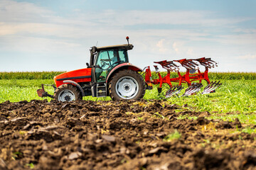 Red tractor is engaged in plowing the soil in a vast green farmland under a cloudy sky