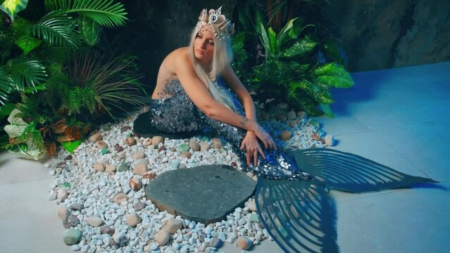 fashion model posing fantasy woman wet mermaid sitting under rain shower bathroom. Sexy beauty blond hair girl sea queen goddess. Creative silver costume fairy river Nymph. Backdrop green plants room
