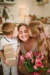 Little son and daughter hugging and kissing mom and make surprise for Mothers Day. Children give bouquet of tulip flowers. Childs gives present box congratulates surprised mom in kitchen. Happy family