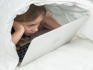 Portrait of a cute little girl using a laptop while lying under a blanket. 