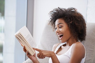 Happy, black woman and reading with book on sofa for novel, story or literature in living room at...