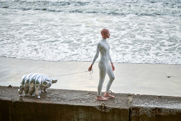 Young hairless girl with alopecia in white futuristic suit walking on concrete fence with toy...