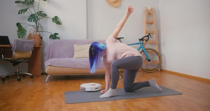 Young plus size latin hispanic teenager girl doing stretching when vacuum cleaner robot working around. Cozy home interior with indoor plants. Online fitness, urban jungle concept