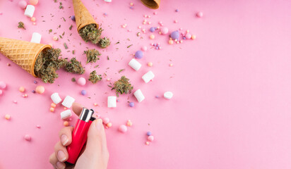 Dry buds of medical marijuana lie in waffle ice cream cones, a female hand holds a lighter on a pink background. There are candies and marshmallows all around. Alternative Cannabis Treatment - obrazy, fototapety, plakaty