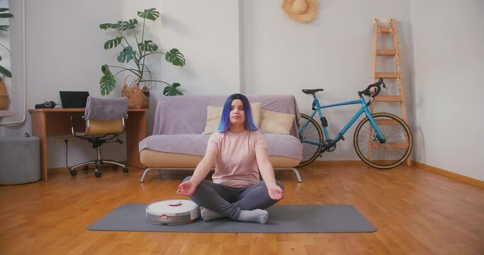 Young plus size latin hispanic teenager girl trying to meditate when vacuum cleaner robot working around. Cozy home interior with indoor plants. Online fitness, urban jungle concept