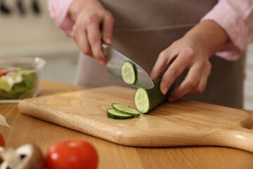 Woman cutting cucumber at wooden table in kitchen, closeup