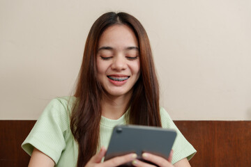 Enthusiastic Asian female student grins while using tablet, seated at  work desk in home environment. Cheerful young learner engaged with her digital pad, comfortably working from her home study space