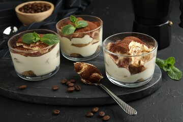 Delicious tiramisu in glasses, spoon, coffee beans and mint leaves on black table
