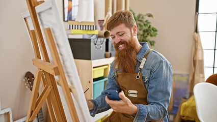 Confident redhead young man, a smiling artist, carefully drawing on smartphone in art studio...