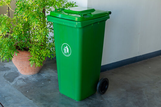 A green plastic wheelie trash bin with a recycling symbol, placed beside a plant pot outside a building.