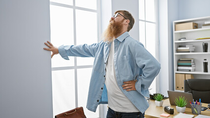 Worried redhead young man, a hard-working office worker, suffering terrible backache while standing...