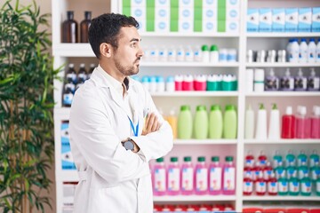 Handsome hispanic man working at pharmacy drugstore looking to the side with arms crossed convinced...
