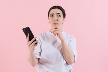 Attractive Caucasian young brunette woman in casual clothes touching chin, thinking and using mobile phone isolated on pink studio background. Technology, work, wireless.