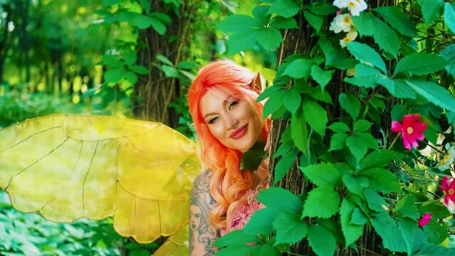 Portrait fantasy happy woman pixie with butterfly wings costume. Girl fairy tale hides behind tree, green leaves forest sun light. Pink dress design red orange color hair pointy elf ears smiling face