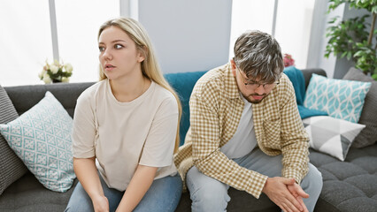 A couple experiencing tension sits apart on a sofa in a modern living room, depicting relationship...