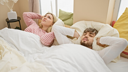 Couple annoyed by loud noise in a bright bedroom, depicting a frustrated woman and a man covering ears.