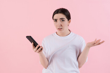 Naklejka premium Confused attractive caucasian young brunette woman in casual clothes uses mobile phone isolated on pink studio background.