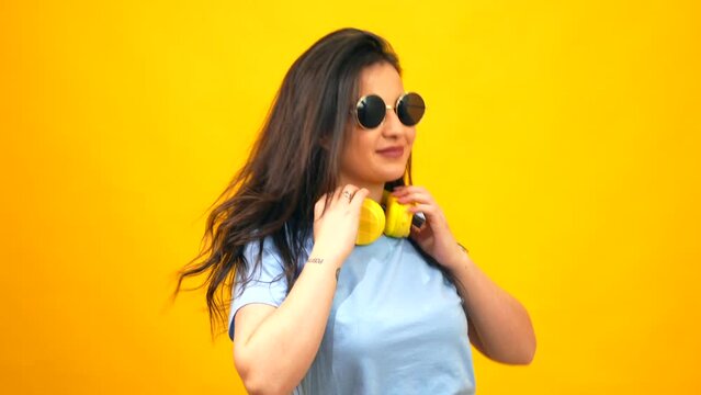 Studio portrait with yellow background of a caucasian woman moving her long hair while holding headphones on neck