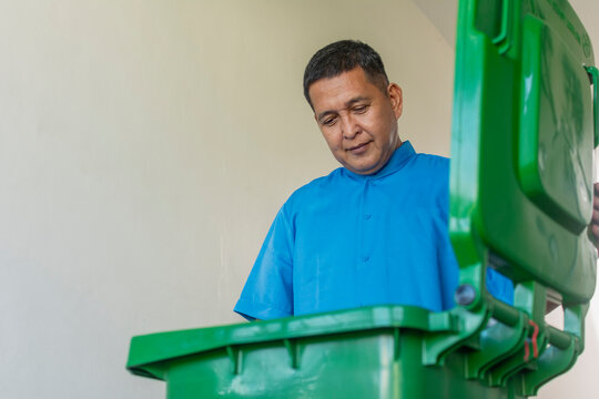 A middle aged asian janitor opens and checks the contents of a large plastic mobile trash bin.