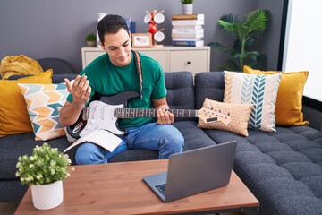 Young hispanic man having online electrical guitar class sitting on sofa at home
