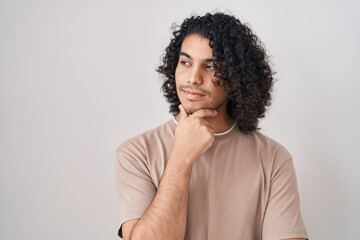 Obraz na płótnie Canvas Hispanic man with curly hair standing over white background looking confident at the camera smiling with crossed arms and hand raised on chin. thinking positive.