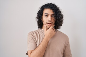 Fototapeta na wymiar Hispanic man with curly hair standing over white background looking fascinated with disbelief, surprise and amazed expression with hands on chin