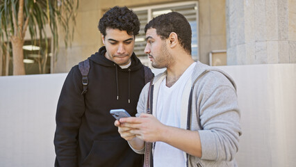 Two hispanic men friends sharing a phone screen together on a sunny city street.