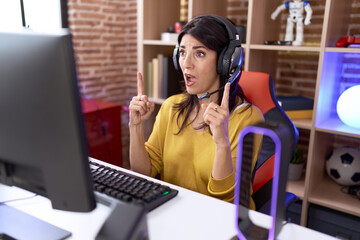 Middle age hispanic woman playing video games using headphones amazed and surprised looking up and pointing with fingers and raised arms.