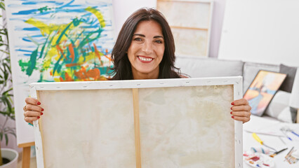 Portrait of a smiling brunette woman holding a blank canvas in an art studio filled with paintings...