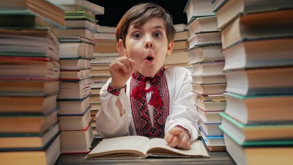 Little ukrainian boy have idea moment, pointing finger up on library books background. Smiling happy pupil child with eureka gesture.