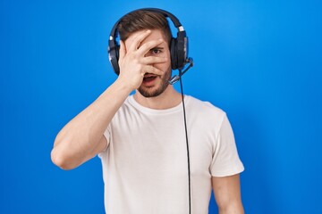 Hispanic man with beard listening to music wearing headphones peeking in shock covering face and eyes with hand, looking through fingers with embarrassed expression.