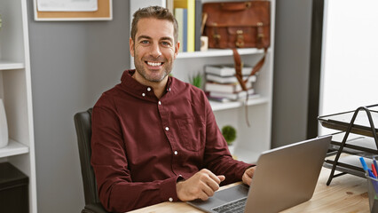 Handsome hispanic man with beard smiles working on laptop in modern office
